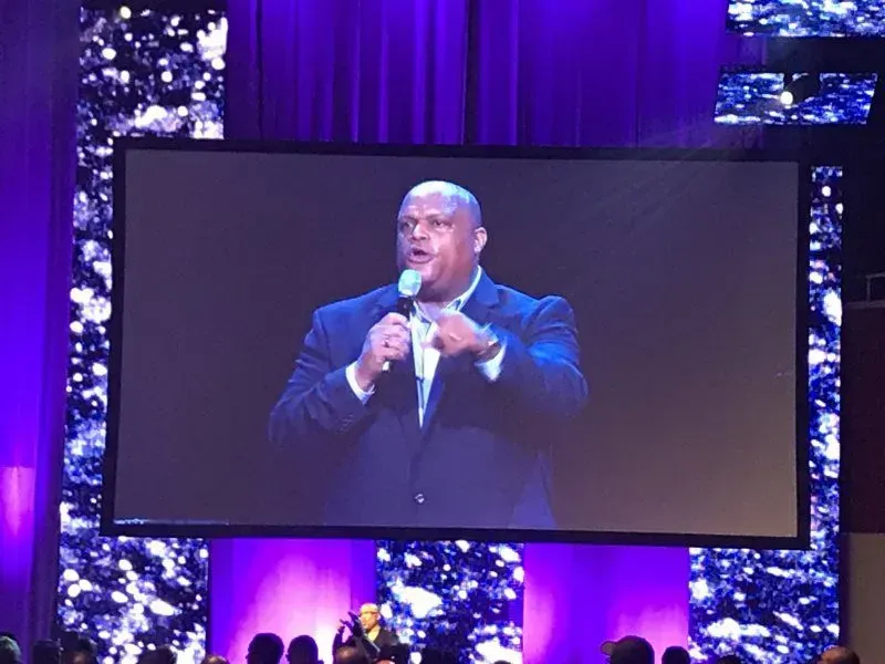 A man in a suit is speaking into a microphone on a large screen.