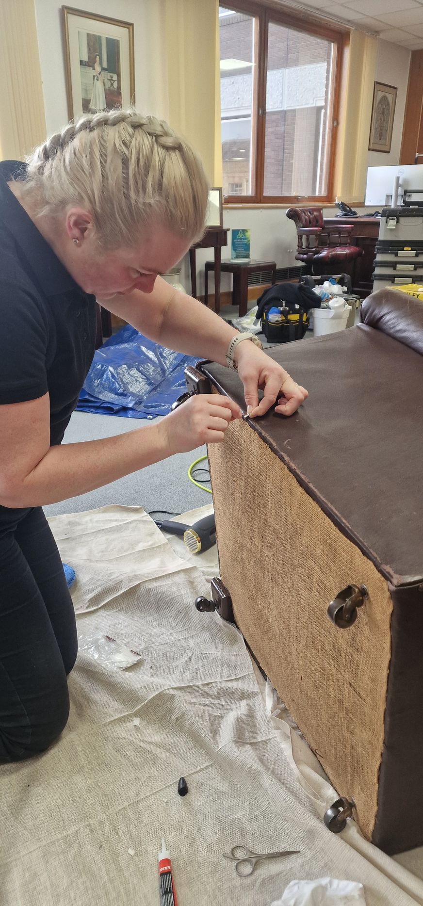 A person is working on a brown furniture piece, possibly upholstery. Kneeling with braided hair.