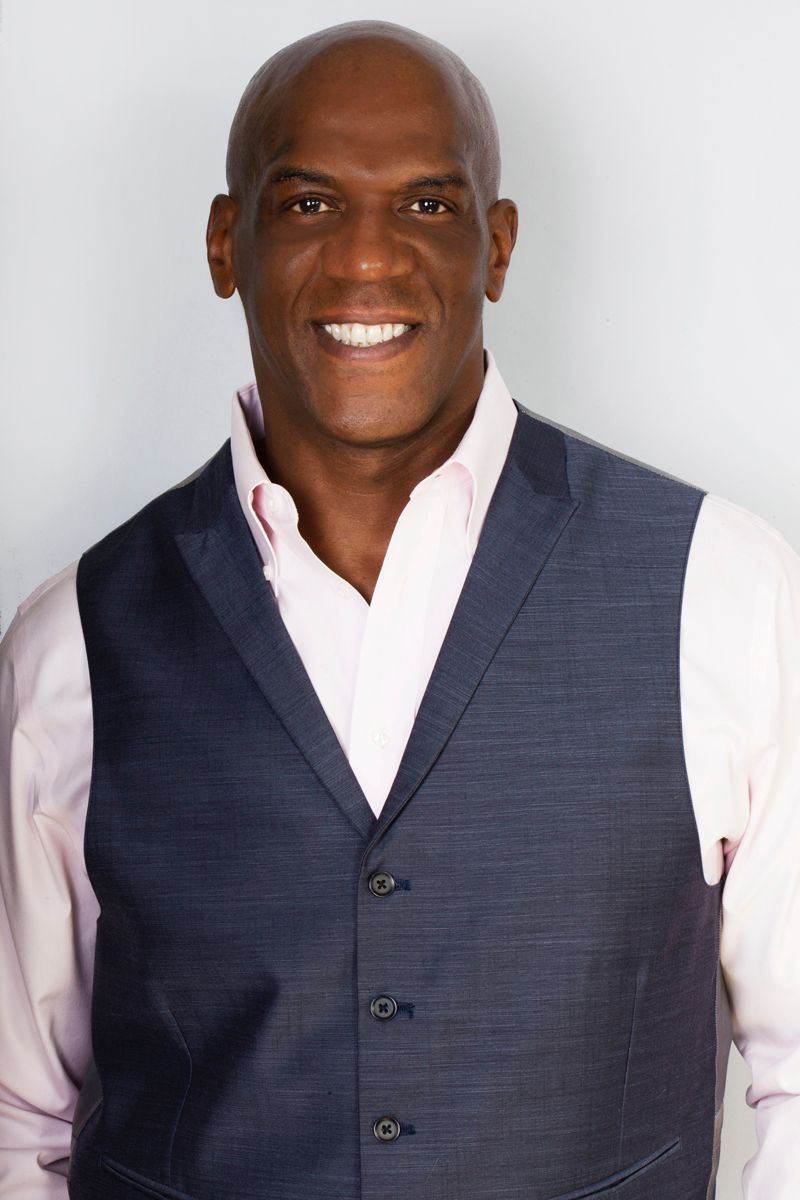 Man in blue blazer and open-collar shirt smiles, set against a golden background.
