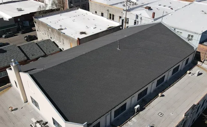 Overhead view of a building with a dark gray roof surrounded by other buildings, a clear sky.