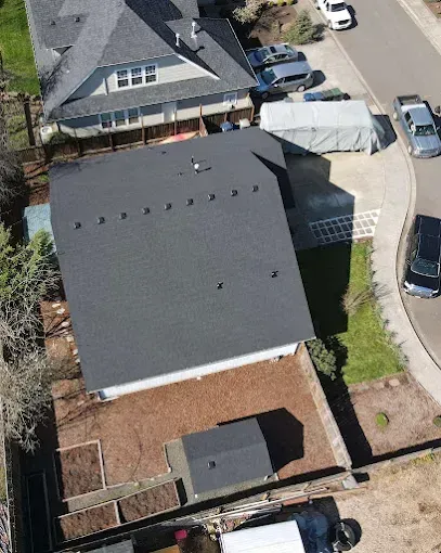 Overhead view of a house with a dark gray roof and surrounding yard. Neighboring houses and cars are visible.