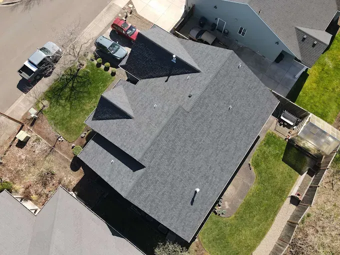 Aerial view of a gray-roofed house with two cars in the driveway, surrounded by green grass and other houses.
