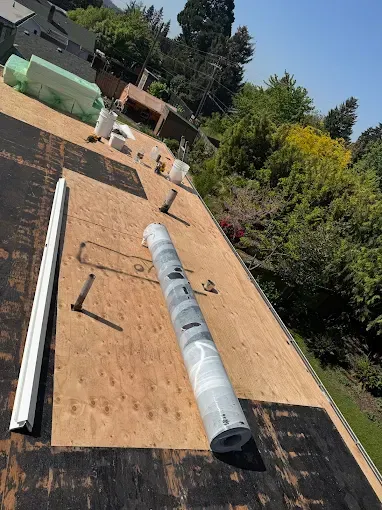 Roofing materials on a partially completed roof; view from above. Plywood, roll of membrane, and other supplies are present.