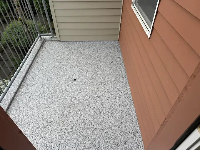 Small balcony with a speckled gray and white floor, brown siding and tan soffit.