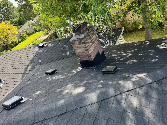 A roof with a brick chimney and vents; the shingles are dark gray.