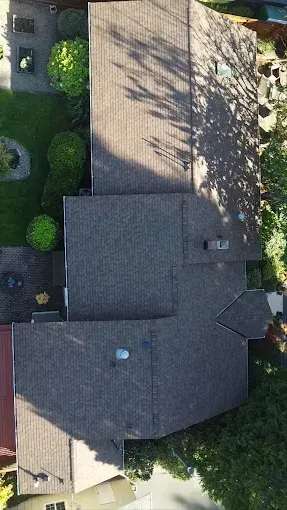 Overhead view of a house with a brown shingle roof, surrounded by green trees and grass, and a small patio.