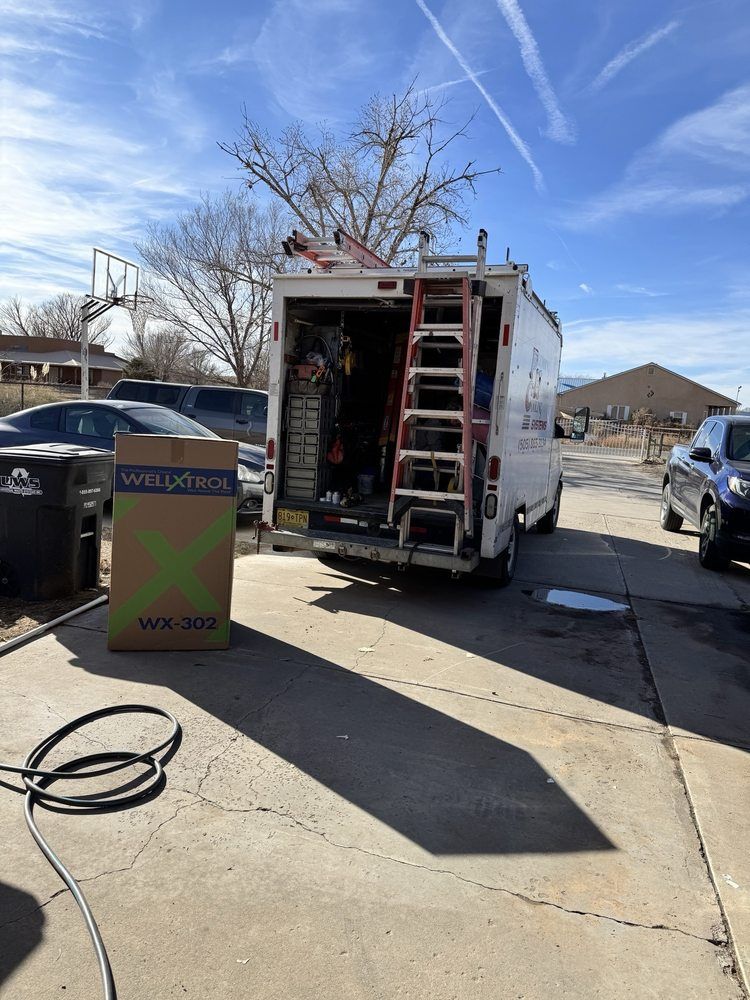 White work van with open back, ladder, tools, and a large box on a driveway.