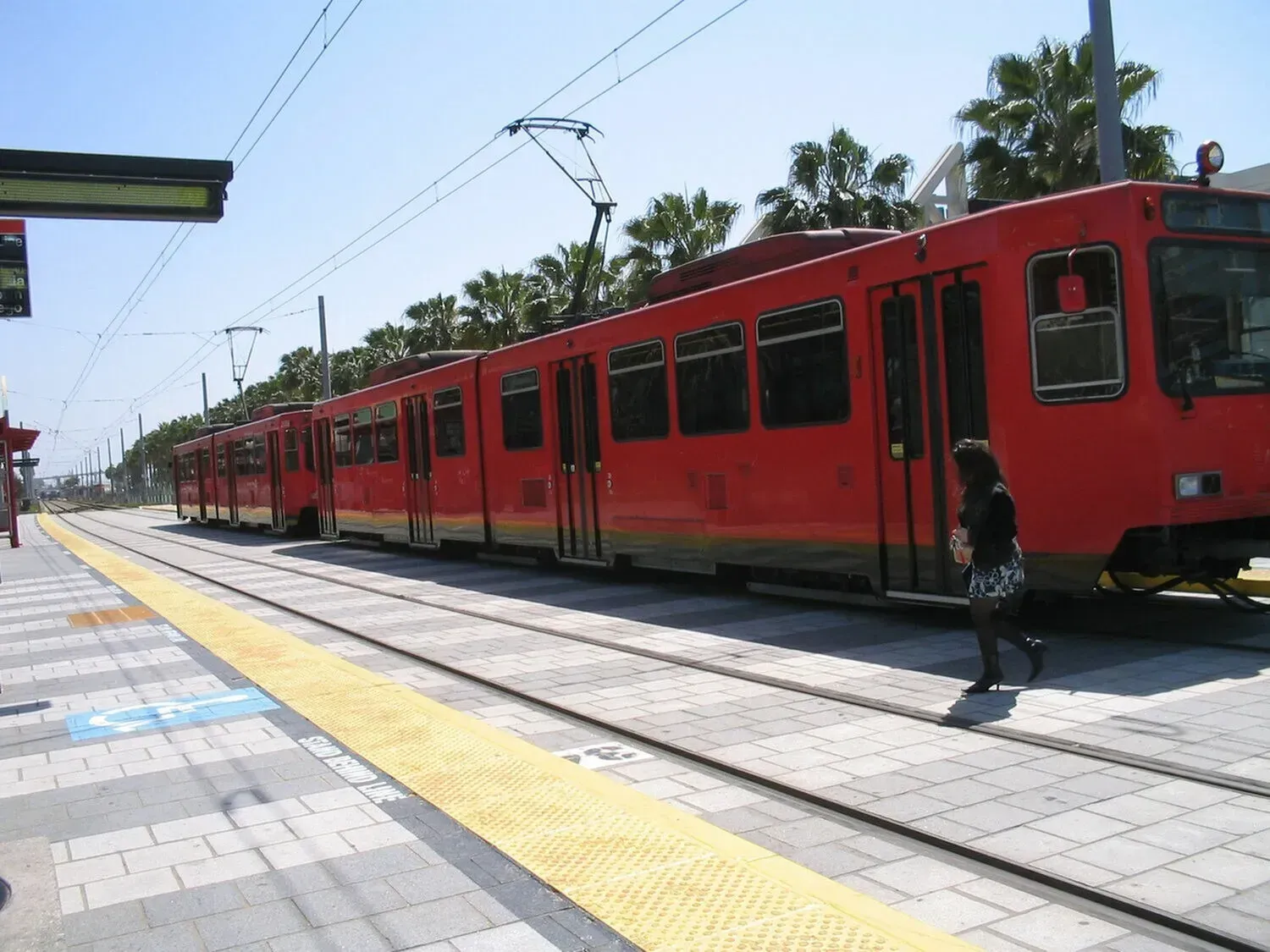 Red trolley car at a station with a person walking across tracks on a sunny day.