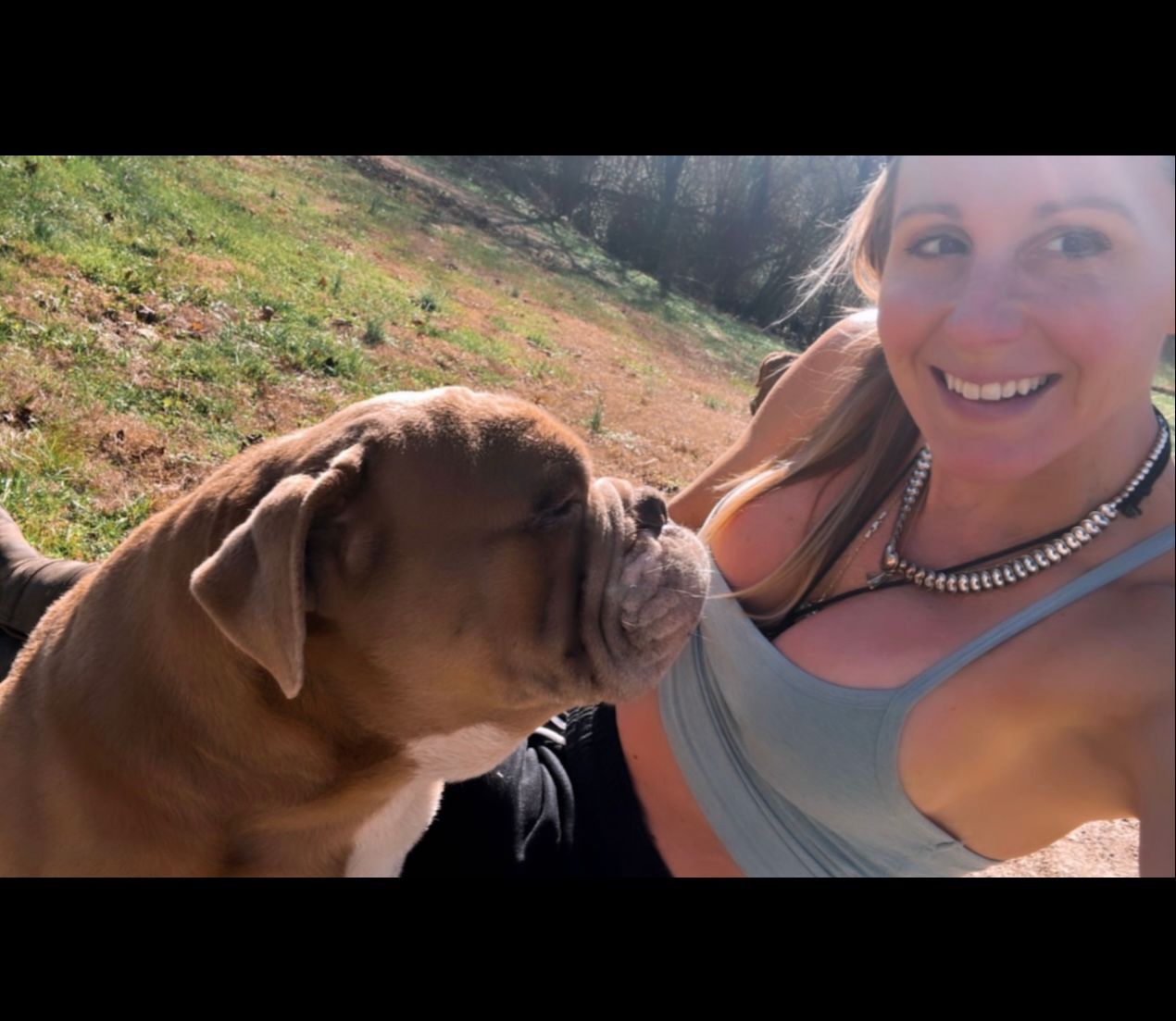 A woman smiles as a brown dog nuzzles her chest outdoors, sunny with grass.