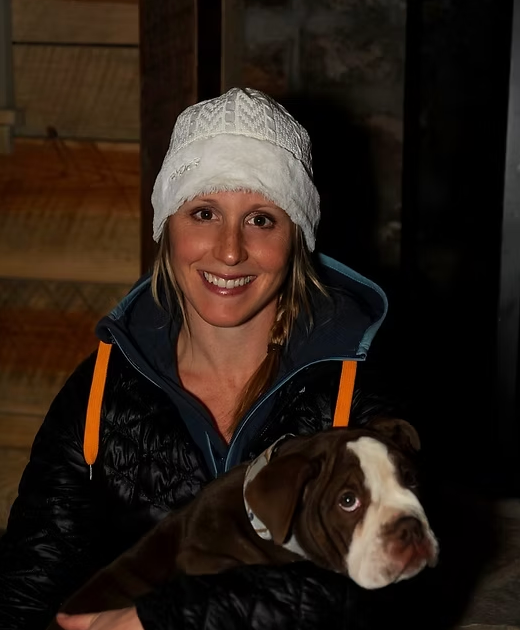 Woman holding a brown and white dog, both smiling. The woman wears a white hat and black jacket.
