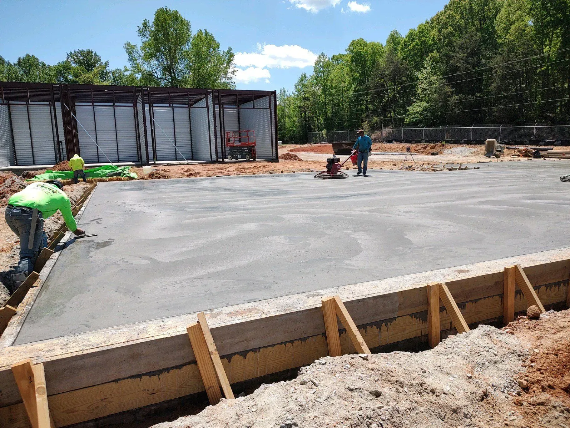 Workers pouring a concrete slab at a construction site with wooden forms and trees in the background