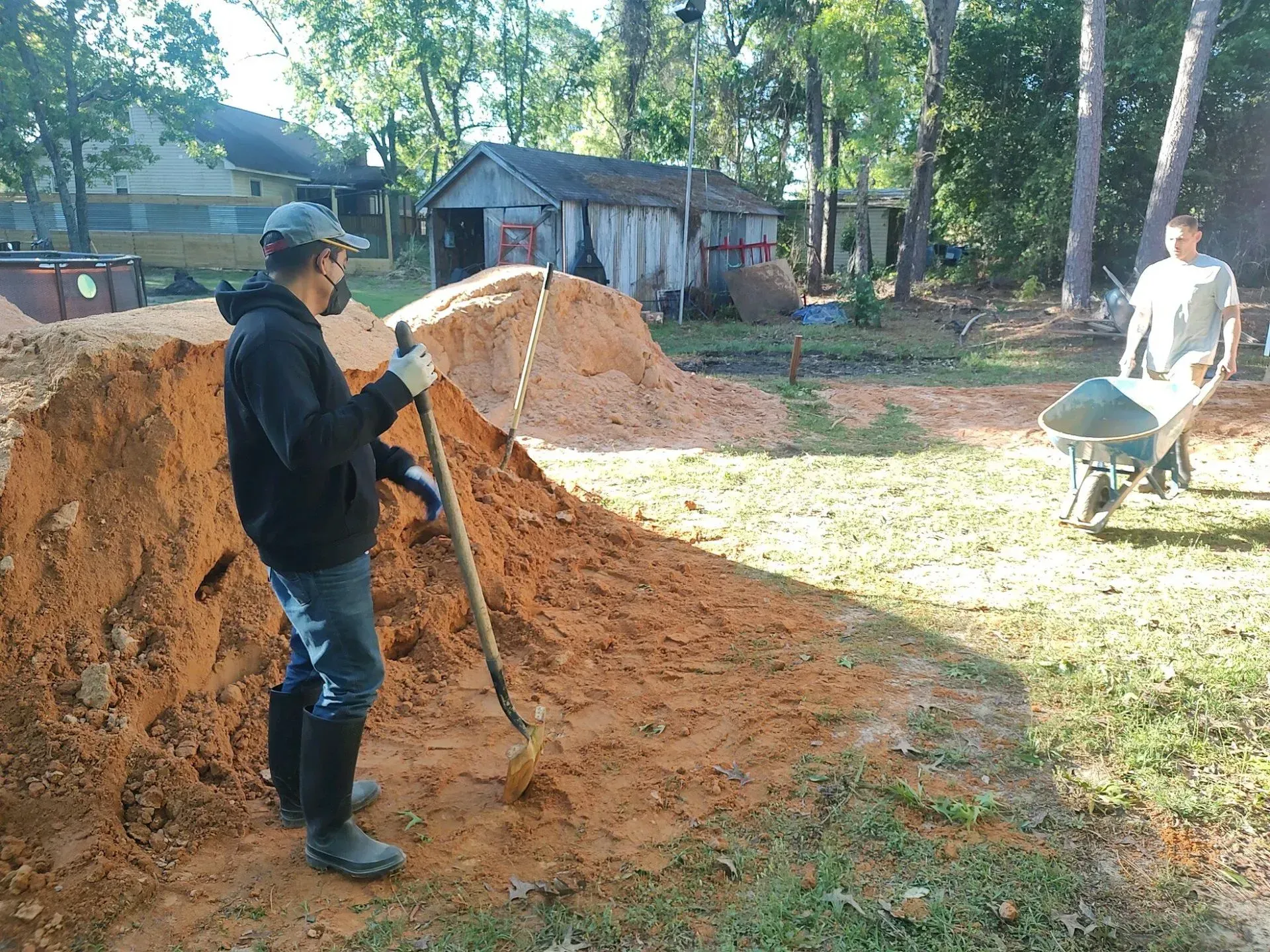 Two workers move dirt with shovels and a wheelbarrow in a yard beside a house and trees.