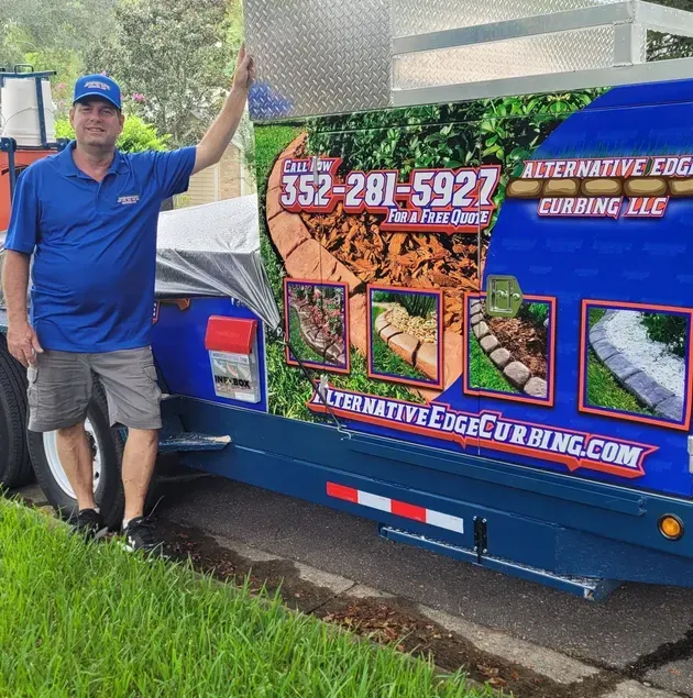Man in blue shirt and shorts standing next to a blue trailer for Alternative Edge Curbing, LLC.
