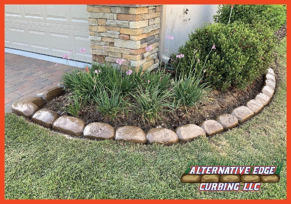 Curved landscaping bed edged with brown stones, containing plants, in front of a house with a stone column.