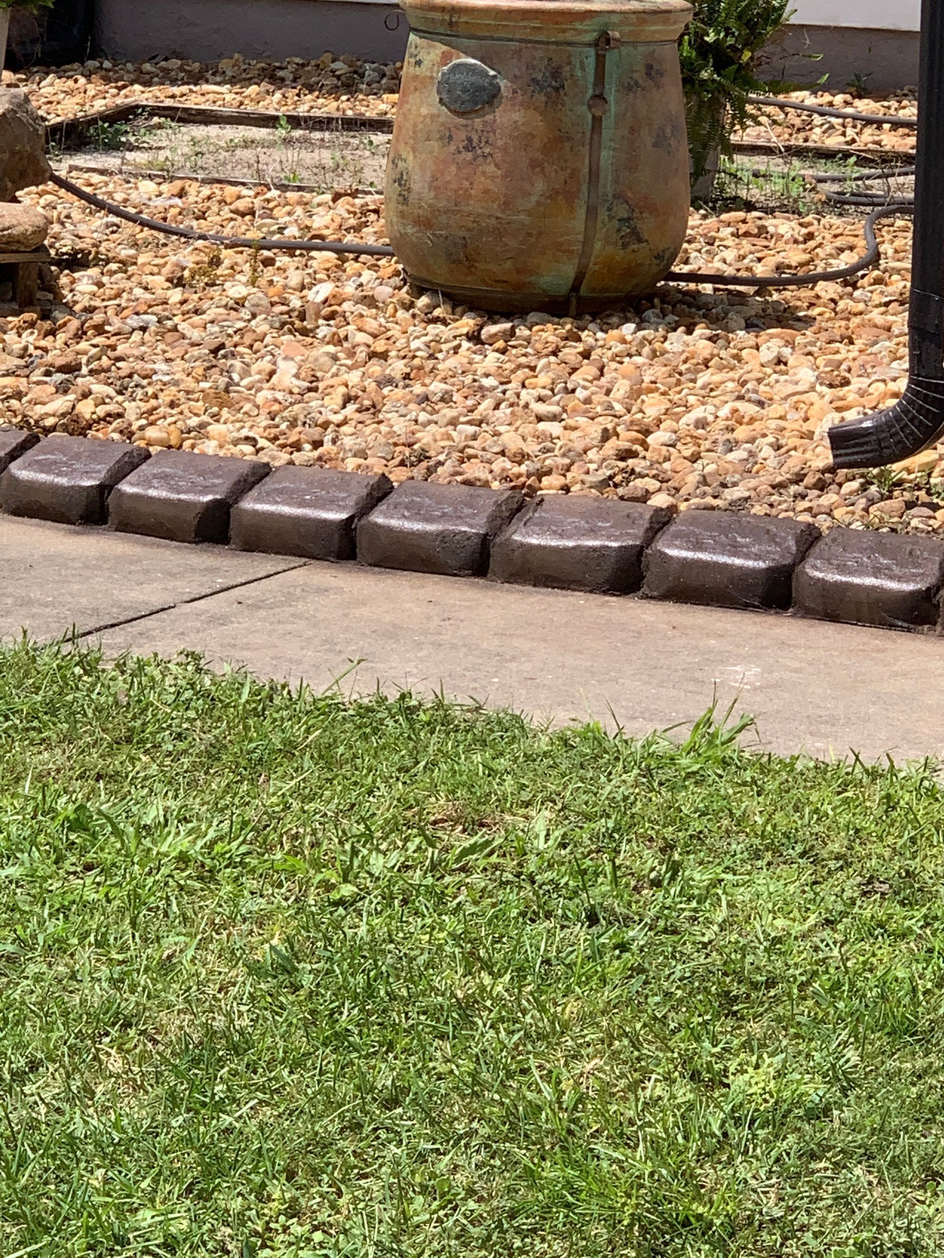 Brown paving stones border a concrete walkway and lawn, with a decorative urn and gravel in the background.