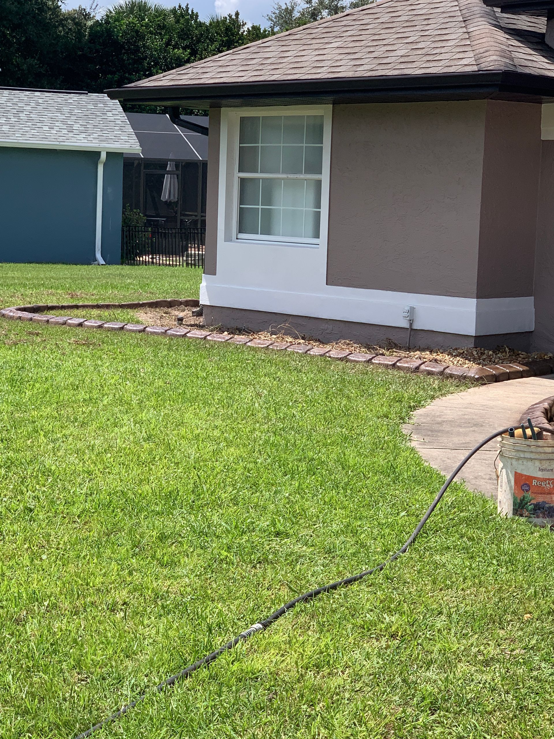 A house with a window, grass, and a garden bed with a black hose on the ground.