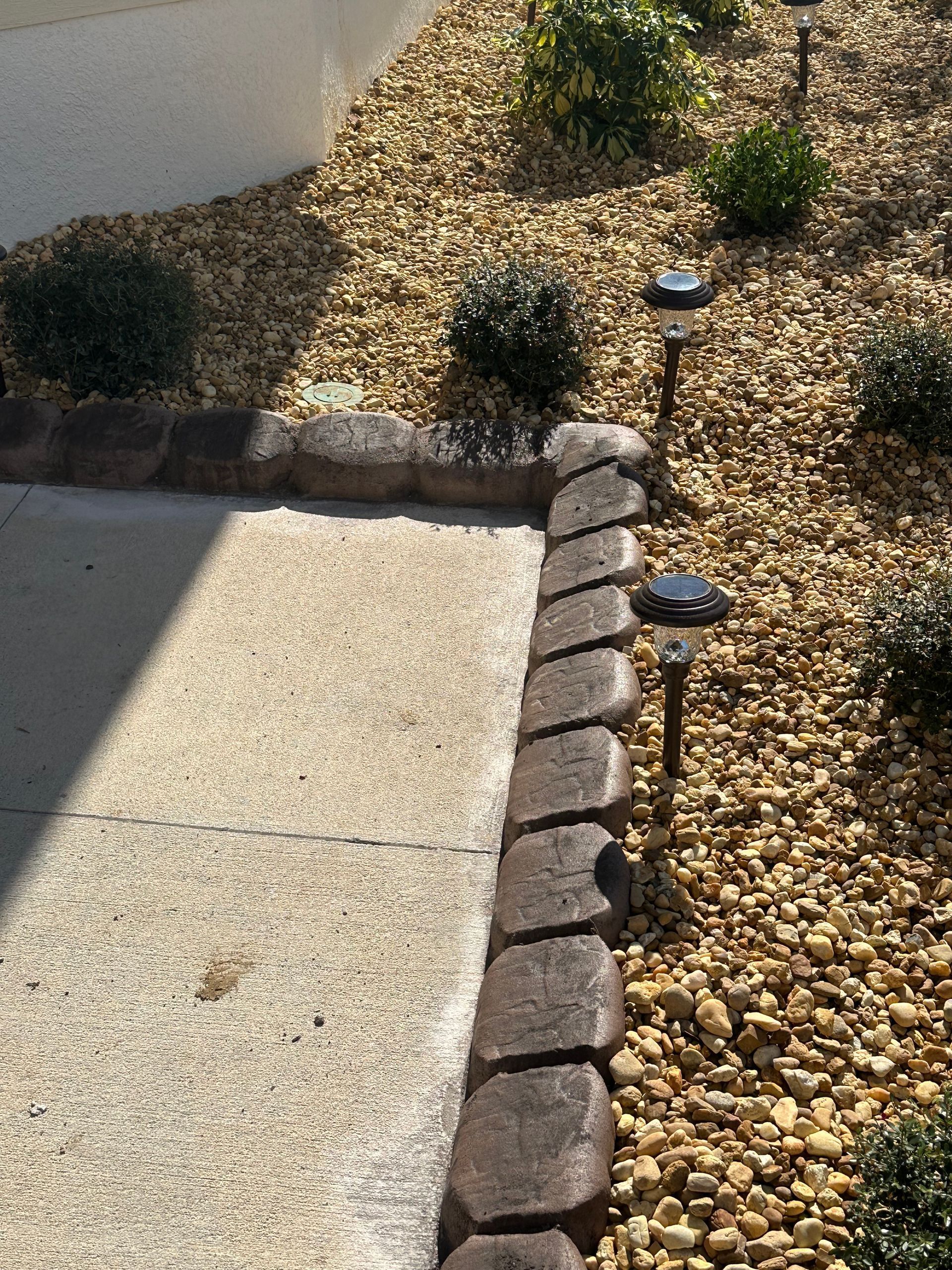 Stone-edged walkway borders a gravel-filled garden with shrubs, solar lights, and a concrete path.
