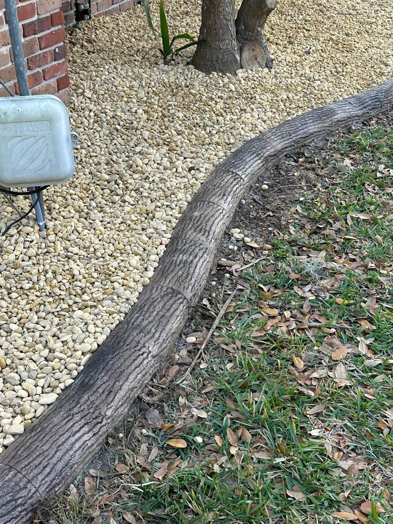 A curved concrete landscape border in a tree trunk design separates gravel from grass near a brick wall.