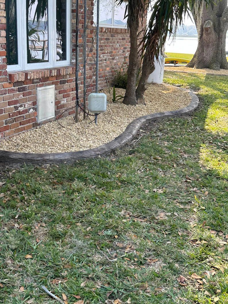 A brick house with concrete landscaping edging, trees, and gravel, beside a grassy lawn.