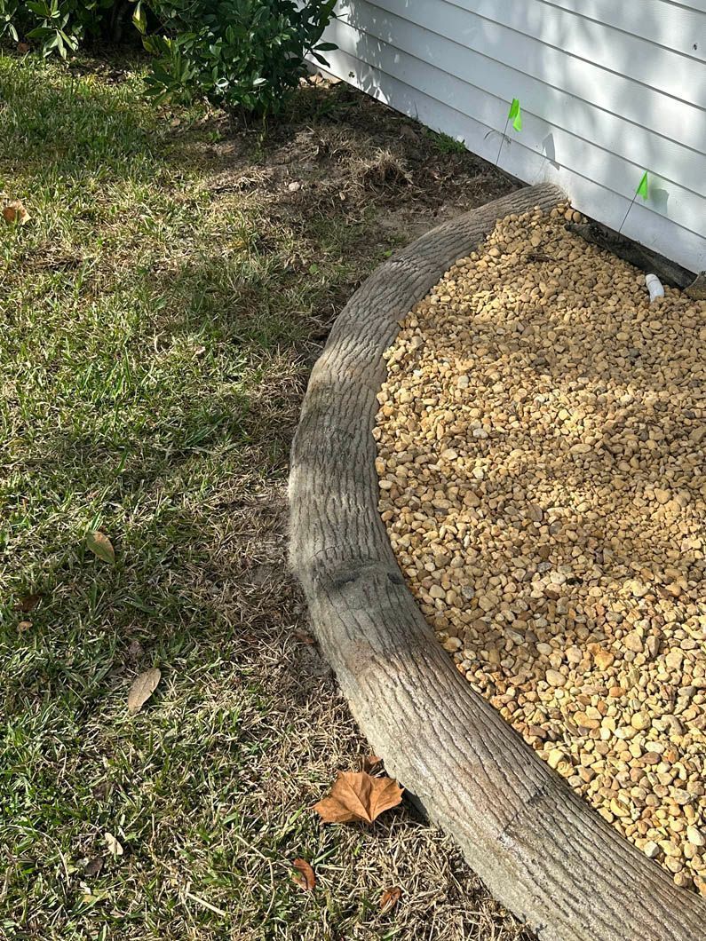 Brown mulch bed bordered by a curved, textured log and grass. White siding.