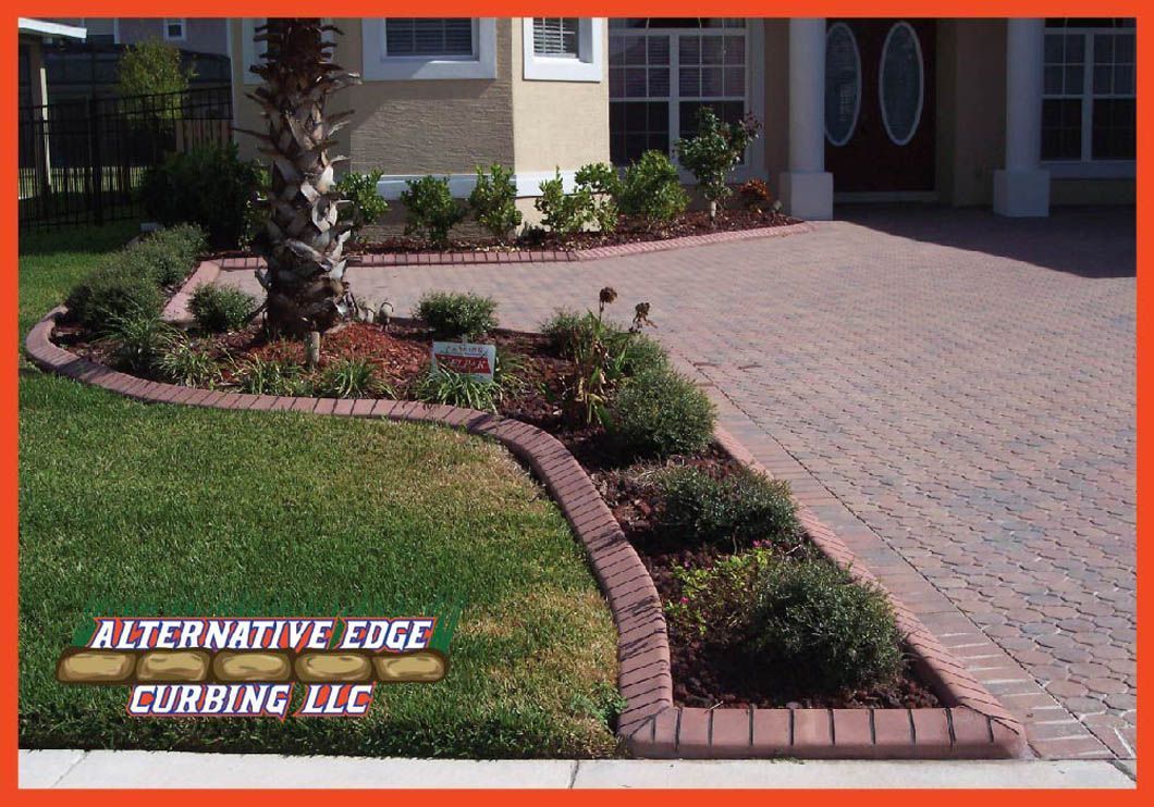 Brick edging separates grass, a flower bed, and a brick driveway in front of a house; logo 