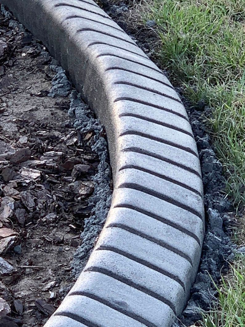 Curved concrete curb with diagonal grooves, bordering dark mulch and grass.