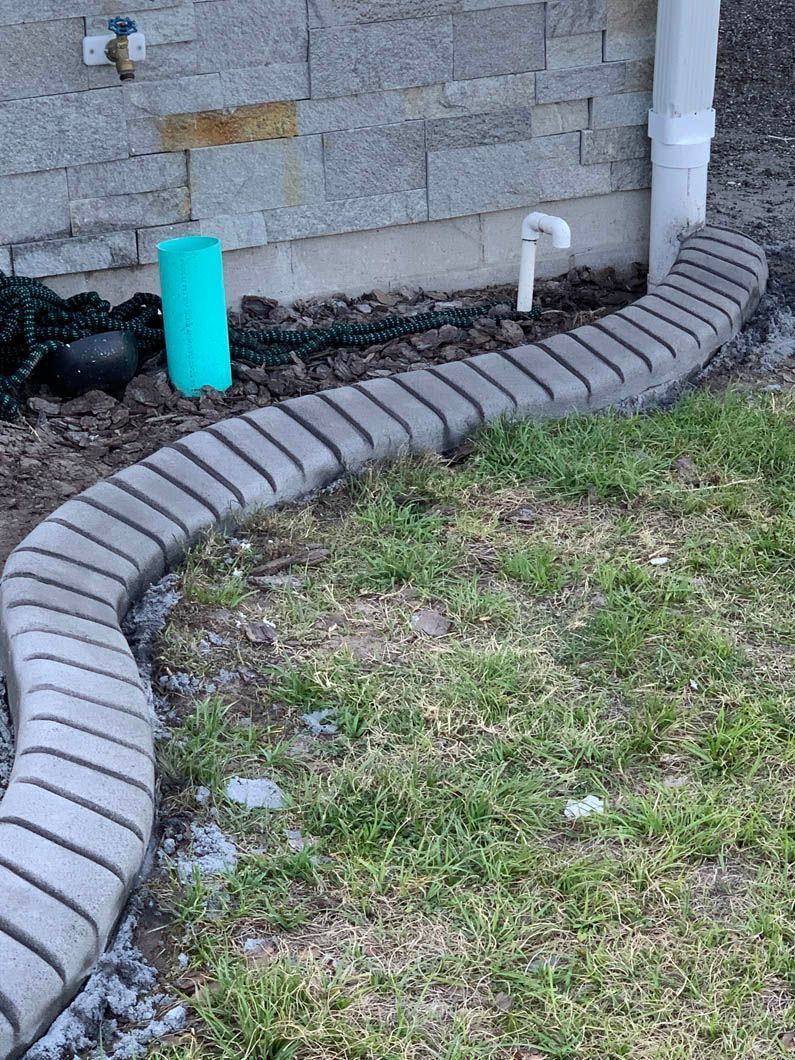 Curved gray brick edging borders grass along a building with a downspout.