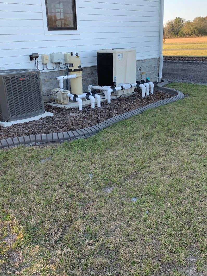 Pool equipment next to a white building, including a heater and filtration system on a concrete pad.
