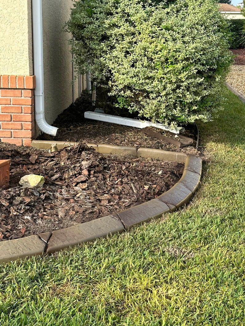 A white downspout drains onto mulch bordered by brown curbing next to a bush and lawn.