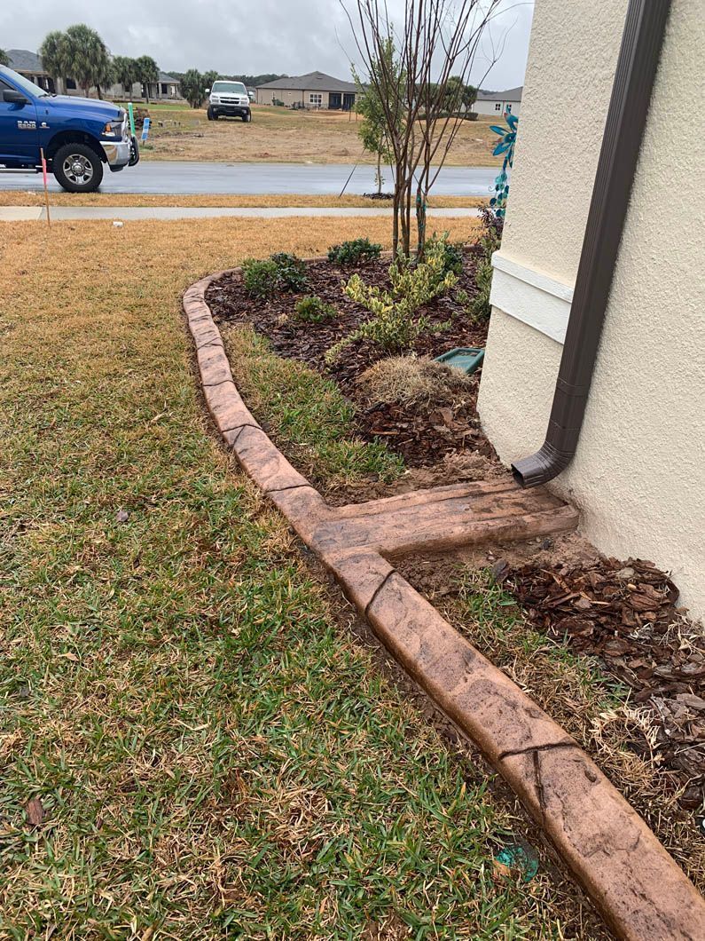 Brown concrete border edging flower bed next to a house. Green grass and a blue truck in the background.