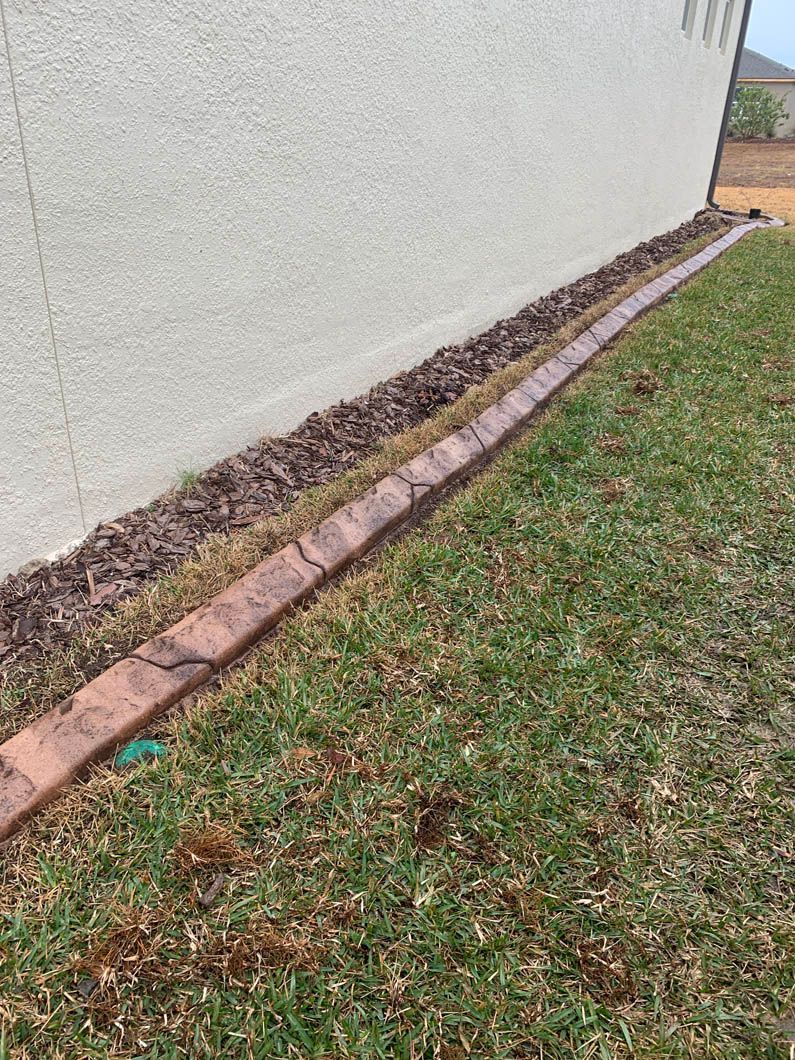 Concrete edging along a beige wall with brown mulch and green grass.