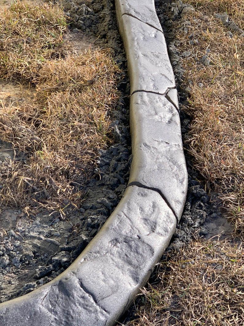 Curved, gray concrete curbing with crack lines separates dry grass from a darker surface.