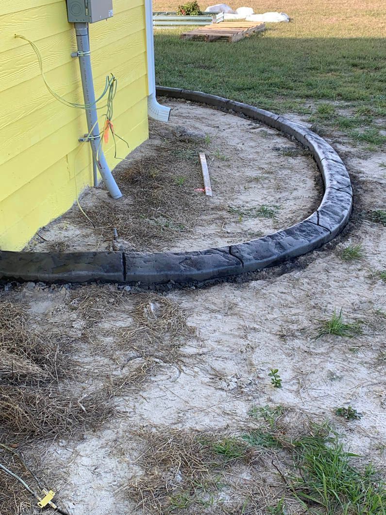 Gray concrete curbing around a yellow house. The curb is set in dirt and dry grass.