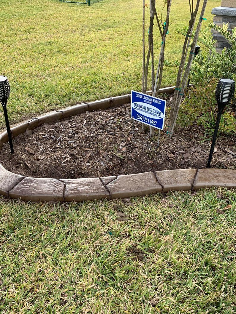 Brown concrete garden edging surrounds a mulched bed with small trees and a sign. Solar lights frame the bed.