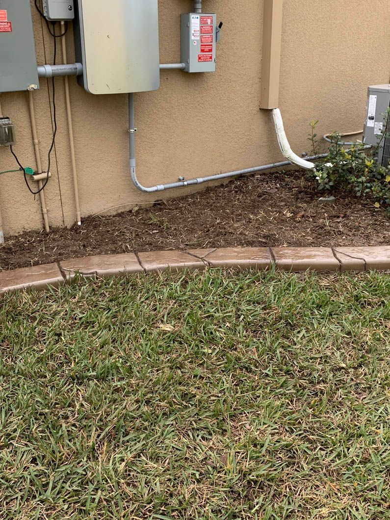 Exterior electrical boxes on a tan stucco wall with a concrete border separating the grass from a mulch bed.