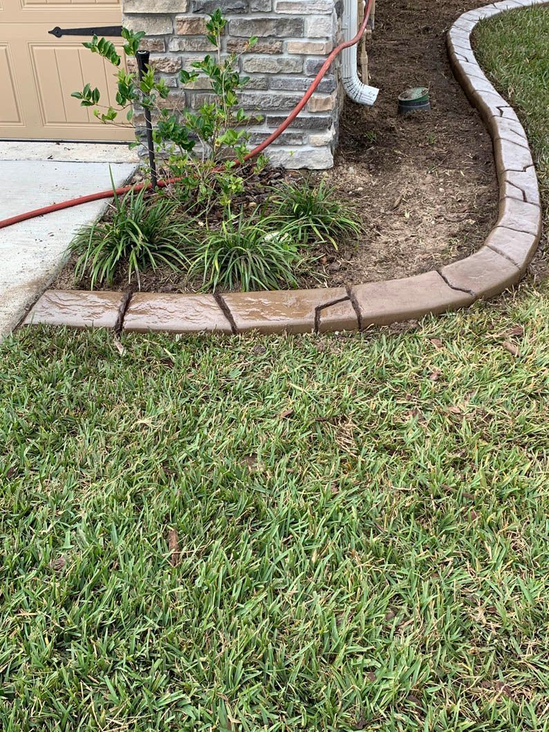 A brown concrete border separates grass from a mulched flowerbed next to a brick house.