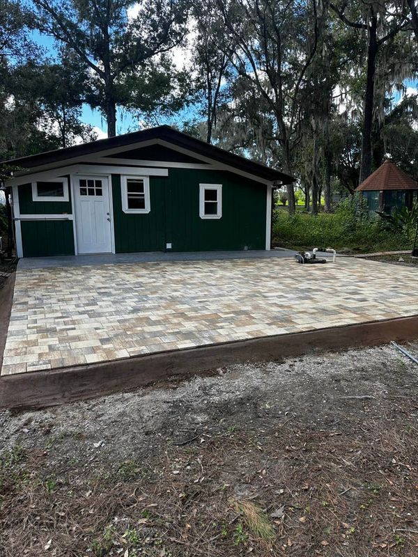 Green building with a white door and brick patio; trees and small gazebo in background.