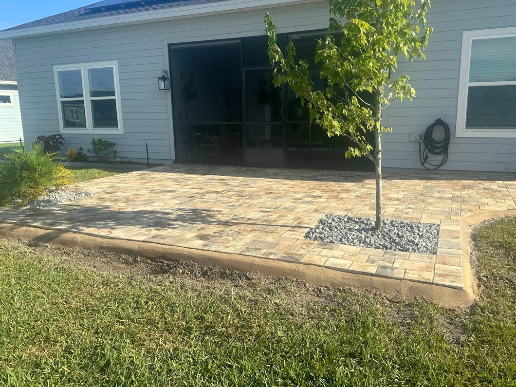 Backyard patio with light-colored pavers, tree, grass, and the back of a house.