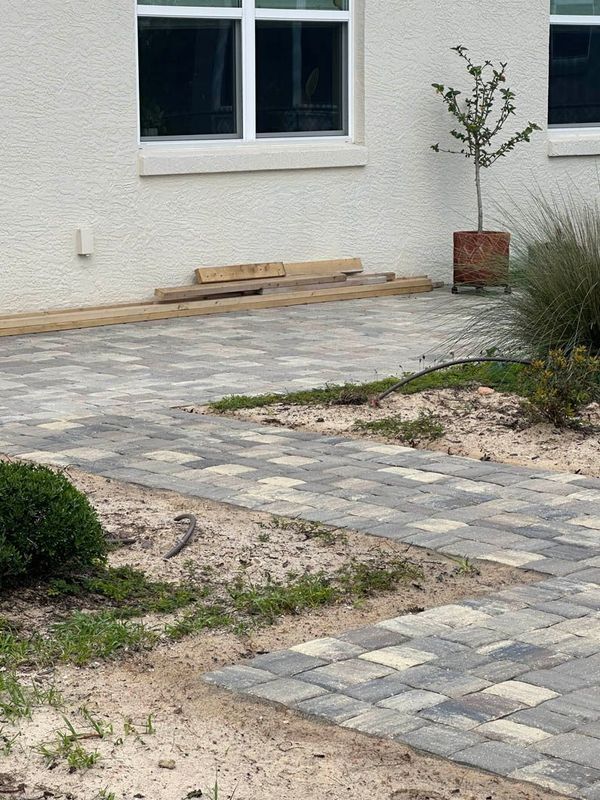 Brick walkway leading to a white building with a window, surrounded by sand and sparse grass.