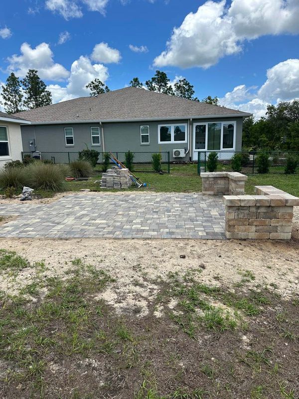 Backyard patio with brick pavers and stone walls, near a light gray house with a blue sky.