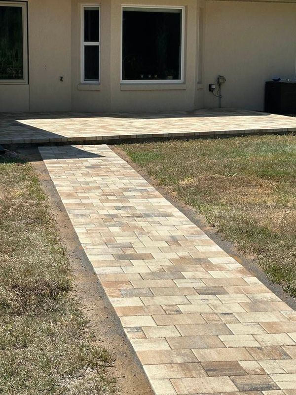 Brick pathway leading from grass to a brick patio outside a light-colored house.