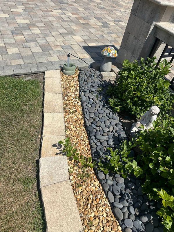 Stone patio with a gravel and rock border beside a garden bed with bushes and a Buddha statue.
