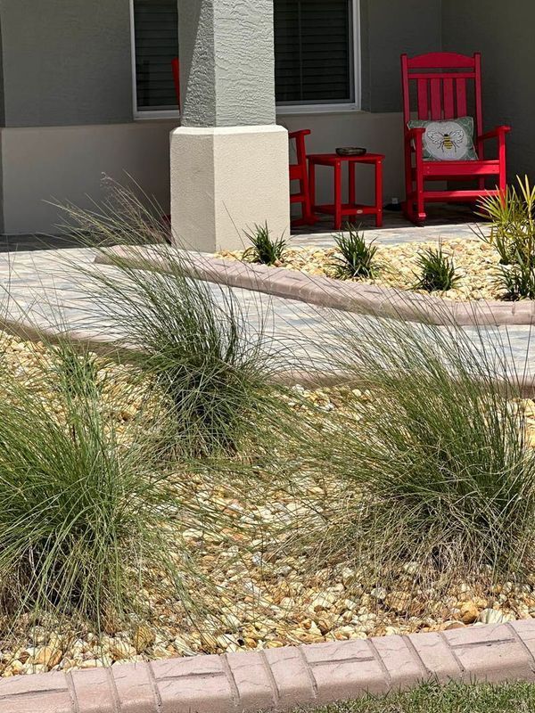 Exterior view with brick border, tan pebbles, green grass, and red rocking chair on porch.