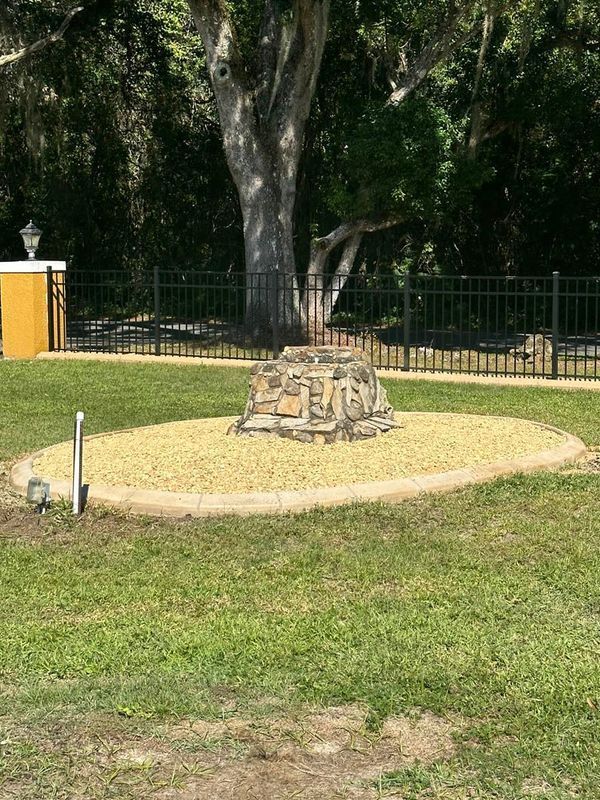 Stone structure in a gravel bed, framed by a black fence and a large tree.