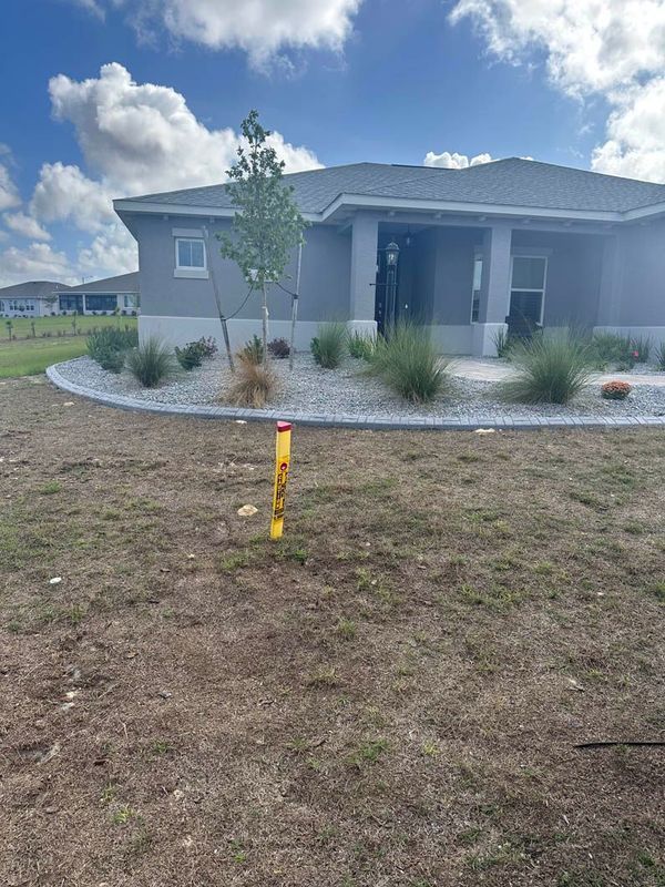 A residential home with a gray exterior, a bare lawn, and landscaping with a utility marker in the foreground.