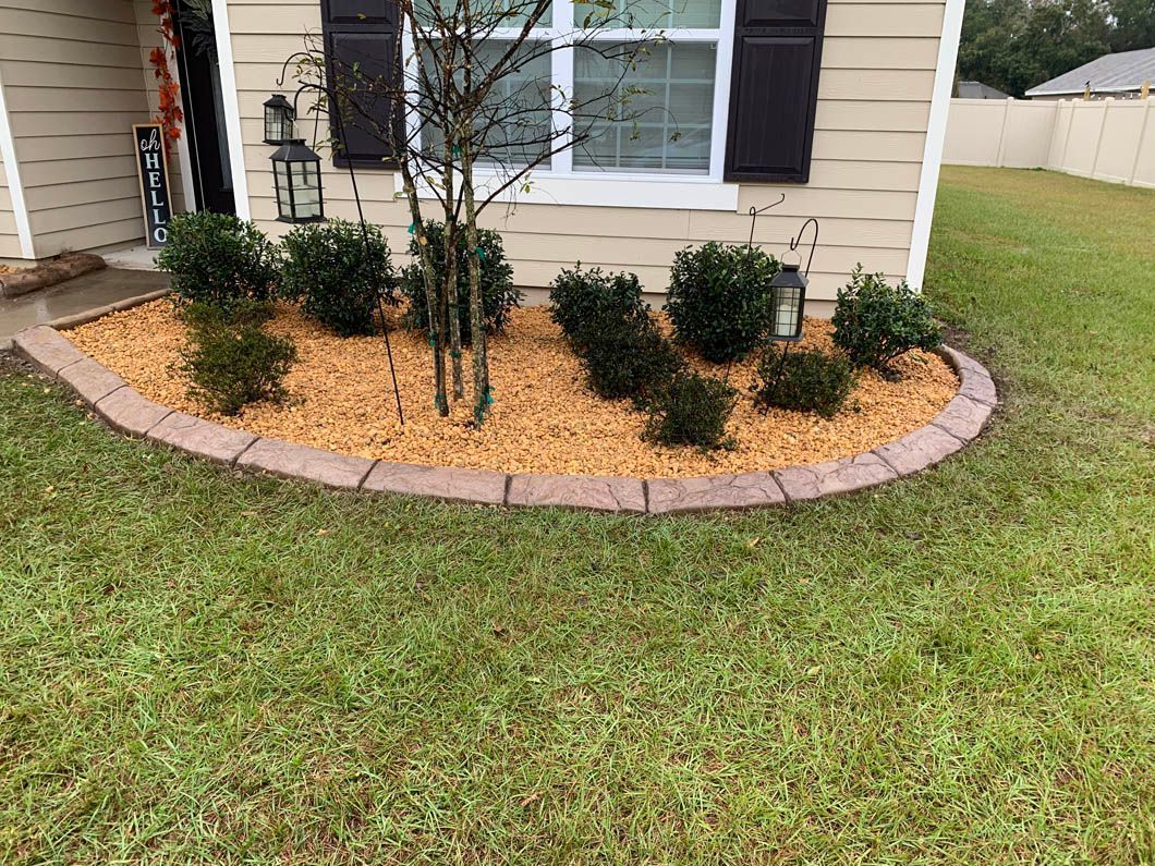 Curved garden bed with brown edging and mulch, green shrubs, and a small tree in front of a beige house.
