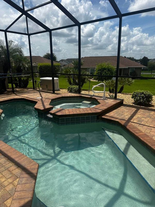 Pool and jacuzzi enclosed by a black screen on a brick patio, with a sunny view of green lawn and houses.