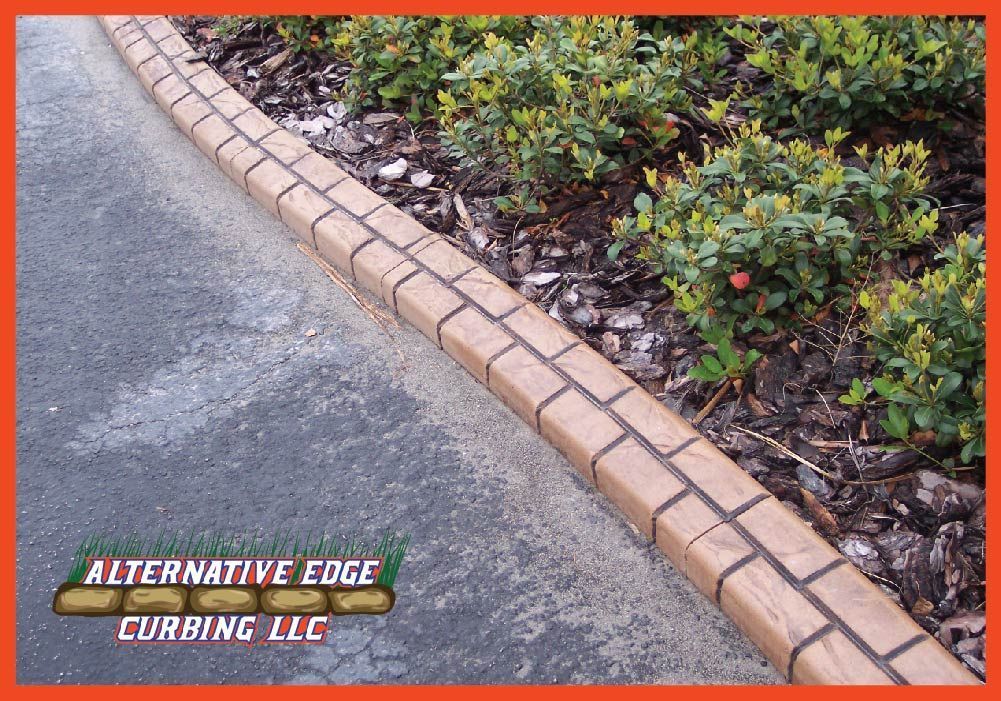Brick-patterned concrete curbing along a paved road separates a landscaped area with green bushes and brown mulch.