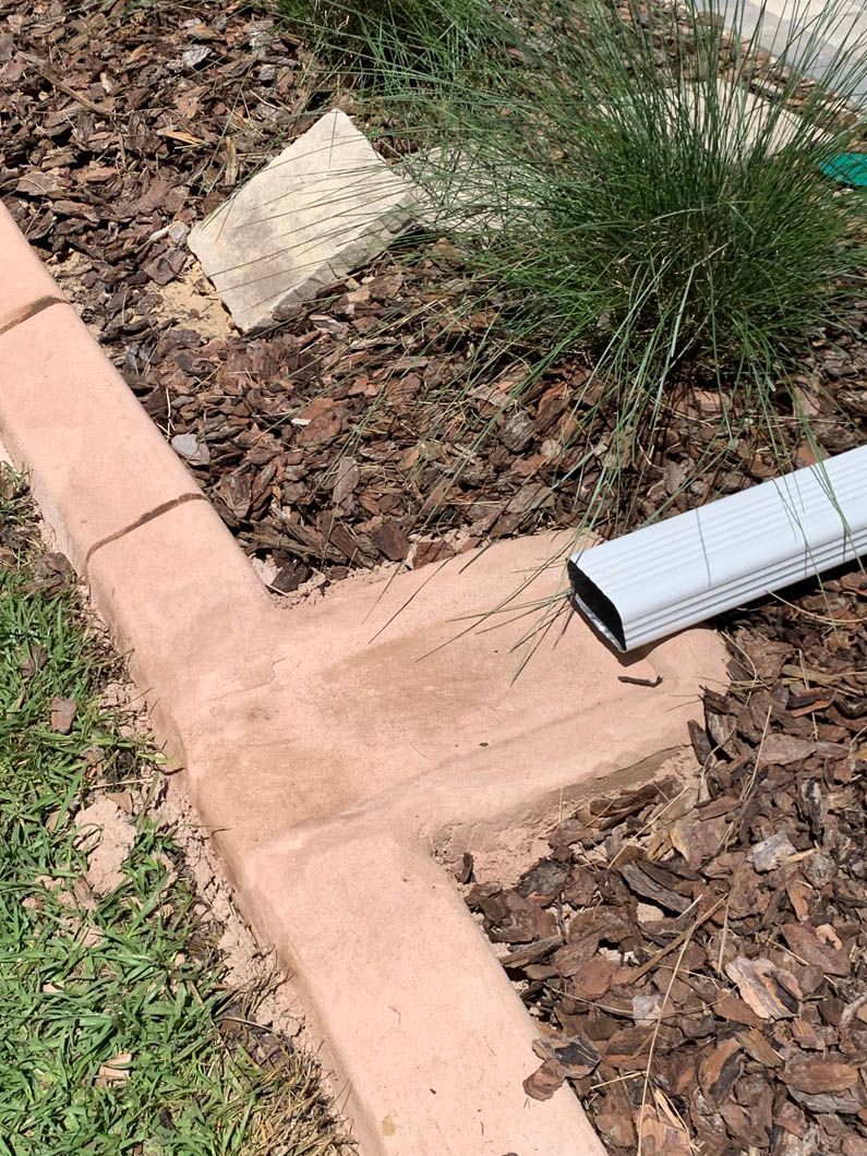 Concrete landscaping border around a garden bed with a drainage pipe; mulch and grass.