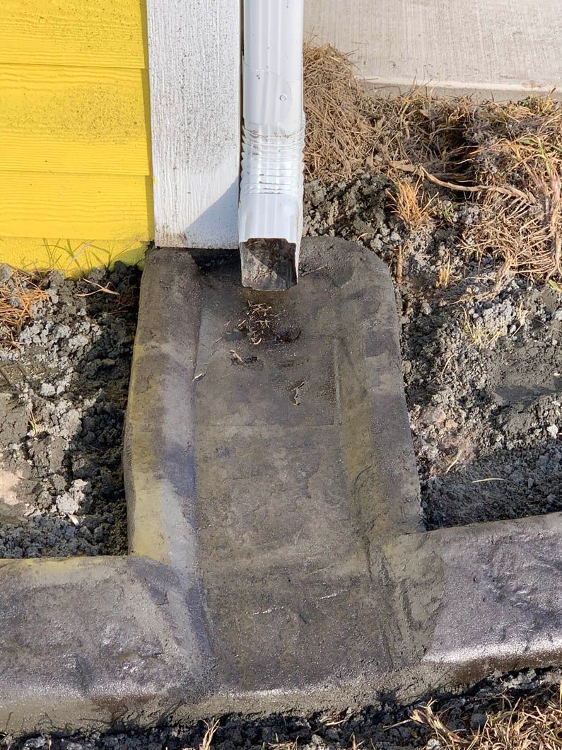 Downspout emptying onto a concrete pad next to a yellow wall and ground.